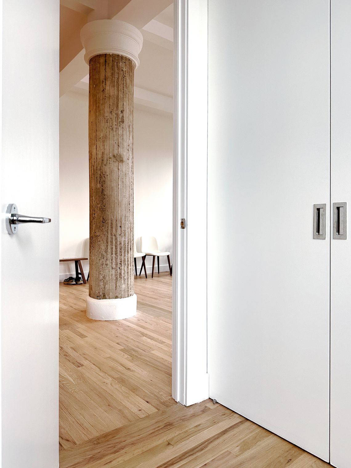 View through a bedroom door of a concrete column, wood floors, and a few chairs in a mostly empty living space.